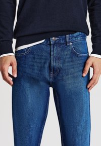 Homme portant un pull bleu marine et un jean bleu avec les mains reposant le long du corps, de la taille au milieu de la cuisse visible.