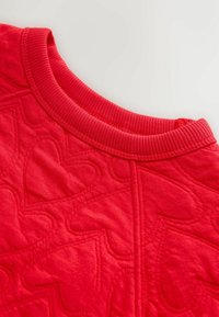 Tissu matelassé rouge avec un motif de cœurs, doté d'un col côtelé. La texture semble douce, avec des accents cousus délimitant les cœurs.