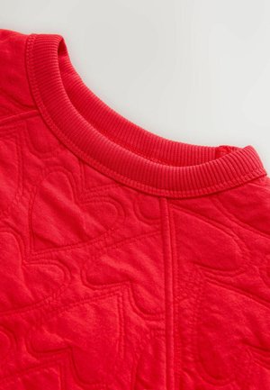 Raudona dygsniuota medžiaga su širdelių raštu, su pintu apykaklės kraštu. Tekstūra atrodo minkšta, su siūtais akcentais, apibrėžiančiais širdeles.