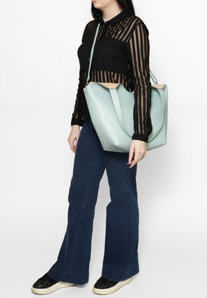 Donna che indossa una camicetta nera trasparente a righe, jeans scuri svasati, sneakers nere e porta una grande borsa celeste a tracolla.