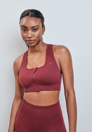 Frau trägt ein ärmelloses, dunkelrotes Sporttop und passende, hoch taillierte Leggings vor einem einfachen hellen Hintergrund.