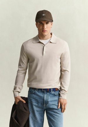 Junger Mann mit beigem Langarm-Poloshirt, blauen Jeans und dunkler Kappe, hält eine dunkle Jacke in einer Hand vor einem schlichten, hellen Hintergrund.