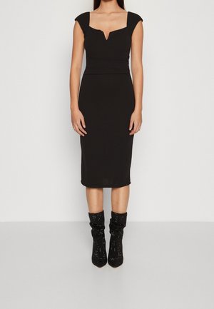 Nainen pukeutuneena istuvaan mustaan mekkoon, jossa on lyhyet hihat ja V-muotoinen pääntie, yhdistettynä mustiin, kimaltaviin polven keskikorkeisiin saappaisiin yksinkertaisella taustalla.