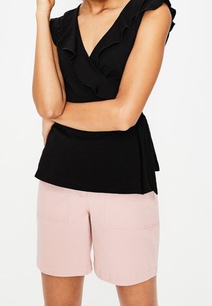 Femme portant un haut noir sans manches à volants et un short rose clair longueur genou, debout les bras croisés.