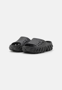 Un par de sandalias negras tipo slip-on con punteras abiertas, parte superior perforada y suelas texturizadas con estrías sobre un fondo blanco.