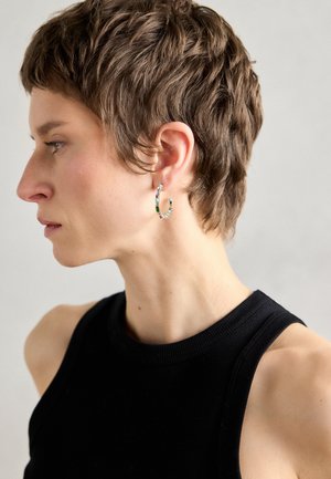 Boucles d'oreilles en argent en forme de hoop ornées de pierres précieuses colorées ; forme circulaire ; portées sur l'oreille externe, contre des cheveux courts et texturés. Haut sans manches noir.