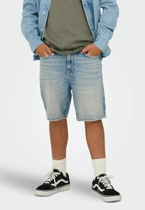 Oseba, oblečena v svetlo modre kratke hlače iz jeansa, zeleno majico, svetlo jeans jakno, bele nogavice in črno-bele superge, stoji z rokami v žepih.