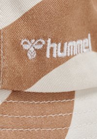 Brun och beige kamouflage-mönstrad keps gjord av texturerad tyg, med en vit broderad "hummel"-logotyp framtill och synliga sömnadsdetaljer.