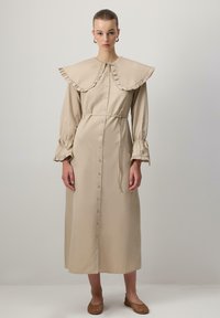 Abito beige a maniche lunghe con colletto arricciato, chiusura con bottoni sul davanti e vita regolata da una cintura. Presenta maniche arricciate e una silhouette dritta.
