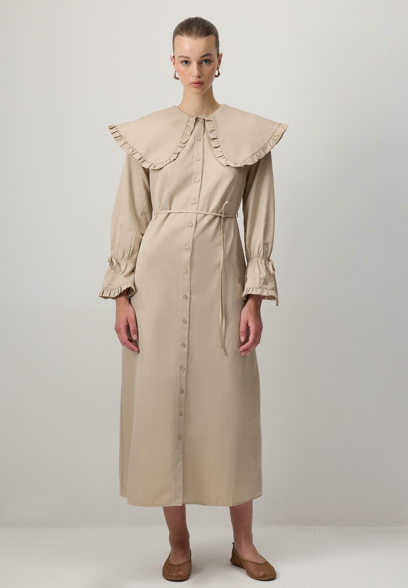 Abito beige a maniche lunghe con colletto arricciato, chiusura con bottoni sul davanti e vita regolata da una cintura. Presenta maniche arricciate e una silhouette dritta.