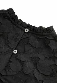 Tissu en dentelle noire avec motifs floraux, deux boutons blancs et un bord froncé le long de la couture supérieure.