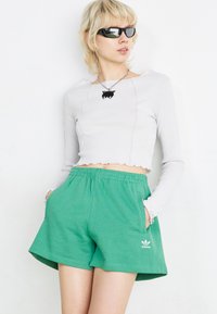 Λευκή ριμπ μπλούζα μακρύ μανίκι με μανίκια και οδοντωτό στρίφωμα, σε συνδυασμό με πράσινες βερμούδες από βαμβακερό μείγμα με πλευρικές τσέπες και λογότυπο Adidas.