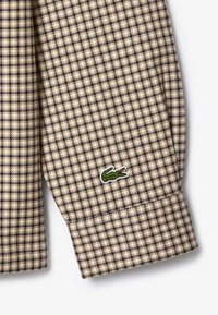 Beżowa i granatowa koszula w kratę z długim rękawem, z wywiniętym mankietem, na którym znajduje się małe, zielone logo krokodyla wyszyte na brzegu.