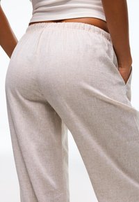 Close-up van een persoon die lichtbeige linnen broek met elastische tailleband draagt en een wit cropped topje, hand in zak tegen een witte achtergrond.