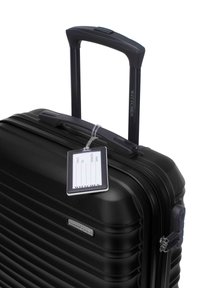 Black hard-shell suitcase with ribbed texture, retractable black handle, and a clear identification tag attached. Features a silver logo plate.