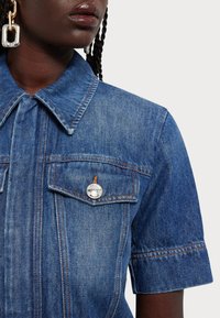 Cămașă din denim de culoare albastru mediu, cu mâneci scurte, două buzunare la piept și un accent cu buton argintiu. Prezintă detalii subtile de cusătură.