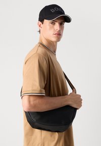 Ung mann i beige t-skjorte og svart caps bærende en svart skulderveske, stående mot en enkel lys bakgrunn, ser inn i kameraet.