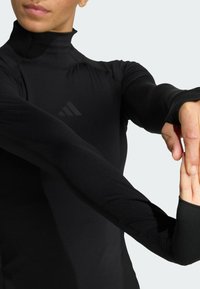 Crni dugi rukavi s visokim ovratnikom i prilagođenim krojem. Ima otvore za palčeve na rukavima i suptilan logo na prsima. Glatka tekstura.