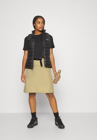 Colete puffer preto sobre uma t-shirt preta, saia bege com um cinto, sapatilhas pretas, segurando uma garrafa de água bege. Fundo simples.