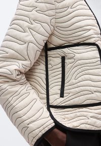 Tuva beigu ķieģeļu krāsas šūta jaka ar melnu abstraktu izšuvumu un rāvējslēdzēja kabatu, ko valkā persona ar tumšu ādas toni.