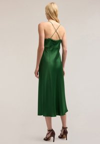 Grön satin midi-klänning med vågig halsringning, korsade axelremmar och slät textur, som når till mitt på vaden och kombineras med bruna klackar.