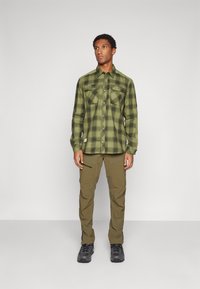 Chemise en flanelle à carreaux verts avec poches poitrine boutonnées, associée à un pantalon cargo olive avec poches zippées et une paire de chaussures noires.