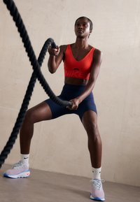 Red sports bra with "NIKE" logo, navy cycling shorts, and multicolored sneakers. The individual is wielding thick black battle ropes.