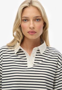 Blonde woman with blue eyes wearing a white and navy striped collared shirt, facing forward against a plain white background.