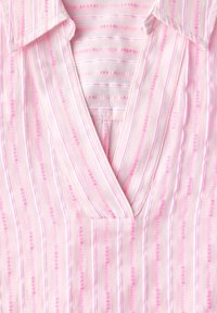 Blouse rose clair avec des rayures verticales blanches et rose foncé, décolleté en V, et un tissu doux et texturé. Manches courtes avec des poignets repliés.