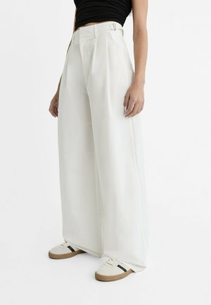 Frau trägt hochtaillierte, weit geschnittene weiße Hose mit Gürtel-Detail, schwarzes Croptop und weiße Sneakers mit schwarzem Streifen.