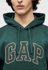 Jeune personne portant un sweat à capuche vert foncé avec de grandes lettres GAP cousues sur la poitrine et des cordons pendant vers le bas.
