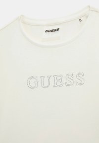 T-shirt en coton blanc avec un col rond et le logo "GUESS" orné de petits clous. Étiquette de taille visible à l'intérieur du col.
