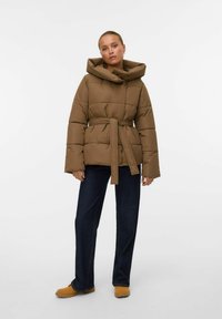 Brun polstret jakke med stor hætte, bæltested og brede ærmer, parret med mørke jeans og brune sko. Glat tekstur og kviltet design.