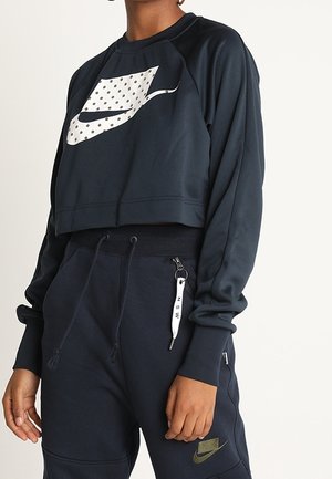 Person trägt ein dunkles, kurzes Nike-Sweatshirt mit gepunktetem Logo und passende dunkle Jogginghose mit Kordelzug und Nike-Logo am Oberschenkel.