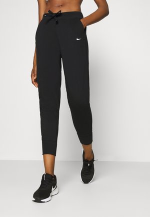 Personne portant un pantalon de jogging noir Nike avec taille à cordon et des chaussures de course noires Nike, debout devant un fond blanc.