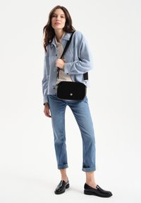 Black suede crossbody bag with a silver button closure, adjustable strap, and minimalistic design. Model wears a light blue jacket and jeans.