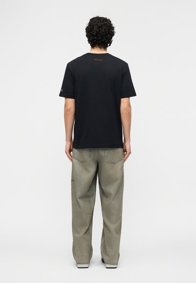 Homme aux cheveux bouclés portant un t-shirt noir à manches courtes avec un petit texte rouge près du col et un jean gris délavé ample, de dos.