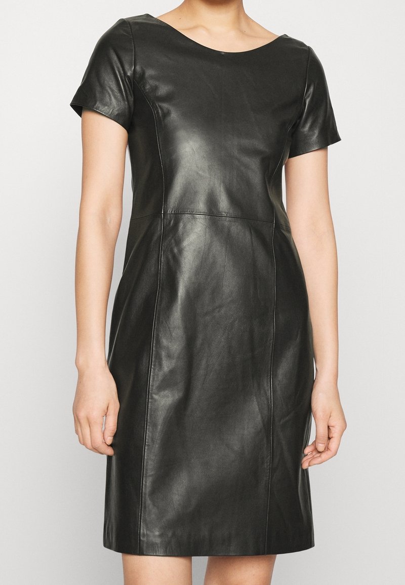 Femme portant une robe courte en cuir noire à manches courtes, taille ajustée et col rond, debout devant un fond clair uni.