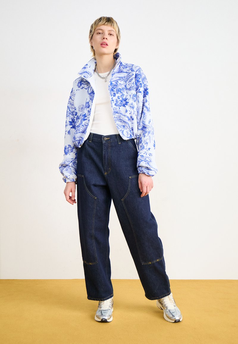Chaqueta con estampado floral azul y blanco, camisa blanca, pantalones anchos de mezclilla oscura y zapatillas plateadas. Ajuste casual y relajado con costuras visibles.