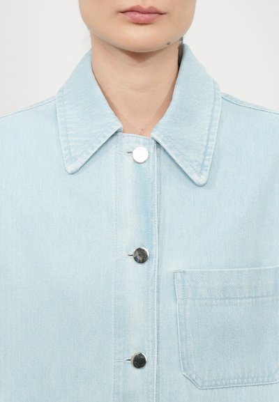 Light blue denim jacket with a spread collar, silver snaps, and a chest pocket. The fabric has a subtle texture and a clean finish.