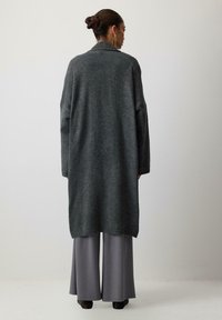 Dlhý, tmavošedý cardigan s voľným strihom, širokými rukávmi a šálovým golierom, kombinovaný so šedými širokými nohavicami, ktoré ukazujú textúrované pletenie.