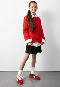 Czerwona sweter z dzianiny z otwartym wzorem, nałożony na białą koszulę z kołnierzykiem. Czarna spódnica plisowana i czerwone trampki z białymi akcentami dopełniają wygląd.