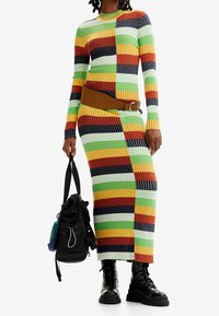 Femme portant une robe en maille côtelée multicolore avec des rayures horizontales et verticales, une large ceinture marron, des bottes de combat noires, et tenant un sac à dos noir.