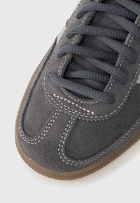 Harmaa mokkanahkainen tennari pyöreillä varpailla, kontrastivärisellä valkoisella tikkauksella, harmailla nauhoilla ja kumipohjalla. Yksinkertainen muotoilu ilman lisäkoristeita.