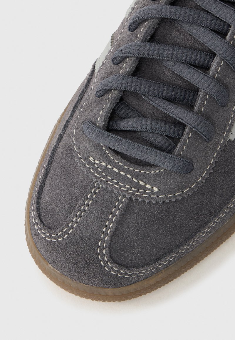 Gray suede sneaker with rounded toe, contrasting white stitching, gray laces, and a gum rubber outsole. Simple design with no additional accents.