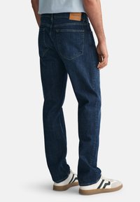 Jeans din denim albastru închis cu croială dreaptă, buzunare din spate vizibile și o etichetă de marcă din piele bej pe talie. Design clasic cu hardware minimal.