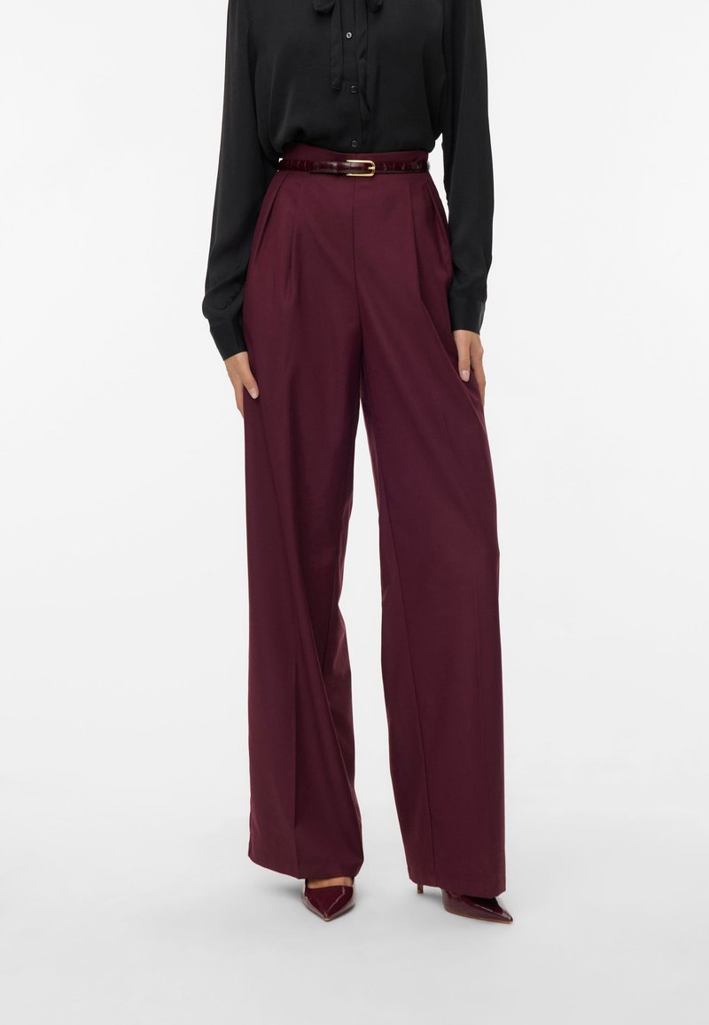 Pantaloni larghi bordeaux in un tessuto morbido con pieghe e vita alta, abbinati a una blusa nera e una cintura coordinata.