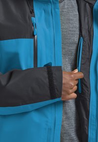 Veste imperméable avec un corps bleu, des manches noires et un intérieur gris. Elle dispose de zippers, de poignets avec velcro et d'une capuche ajustable avec cordon.