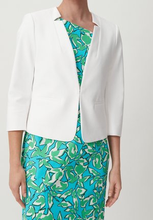 Kvinde iført en hvid cropped blazer over en blå kjole med grønne og hvide abstrakte bladmønstre, stående mod en ensfarvet baggrund.