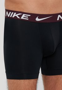 Čierne boxerky s vínovým elastickým pásom, na ktorom je biely nápis "NIKE". Tkanina vyzerá hladko a pružne, poskytuje tesný strih.
